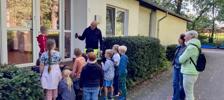 Eine kleine Gruppe von Kindern steht um einen Mann in Handwerkermontur, der ein kaputtes Stück Wasserrohr präsentiert.